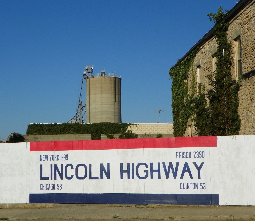 The Lincoln Highway Mileage sign in Franklin Grove is painted on a white picket fence with red and blue. It is 999 miles to New York, 2,330 miles to San Francisco, 93 miles to Chicago, and 53 miles to Clinton, Iowa.